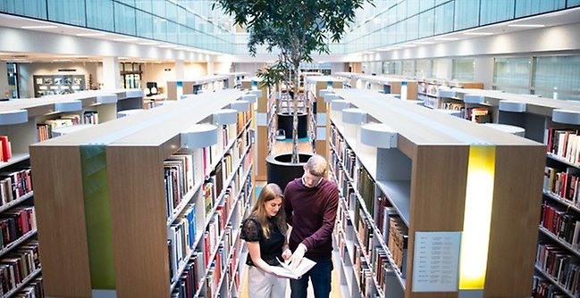 Två studenter som tittar i en bok på ett bibliotek.