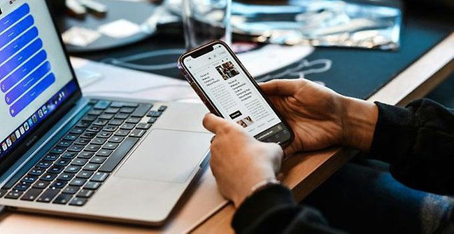 Dator på ett skrivbord samt en mobiltelefon i en persons hand. 